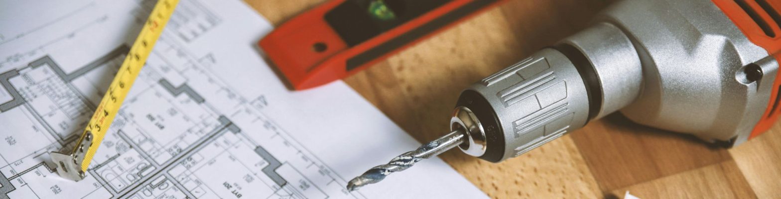 Close-up of a construction workspace featuring a power drill with a metal bit, a yellow measuring tape, a laser level tool, and architectural blueprints spread on a wooden surface.