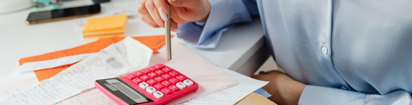 A business professional wearing a light blue blouse sits at a desk, holding a pencil while reviewing financial documents and receipts. A bright red calculator is on the desk alongside orange sticky notes, a mobile phone, and a pen holder filled with office supplies. The scene conveys a focused and organised approach to managing finances or payroll tasks.